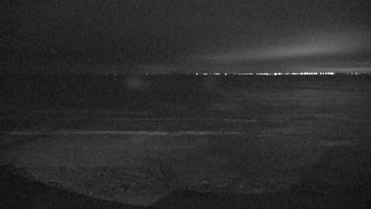 A dark, nighttime beach scene shows a distant line of lights across the water under a mostly clear sky.