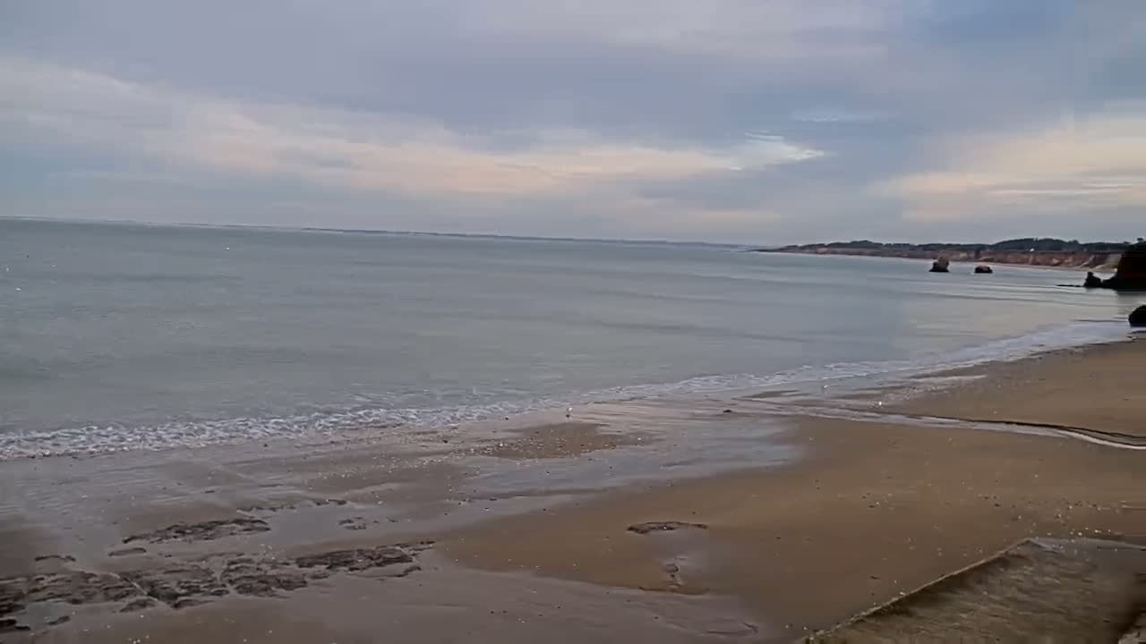 A sandy beach slopes gently down to a calm, gray sea under a cloudy sky.