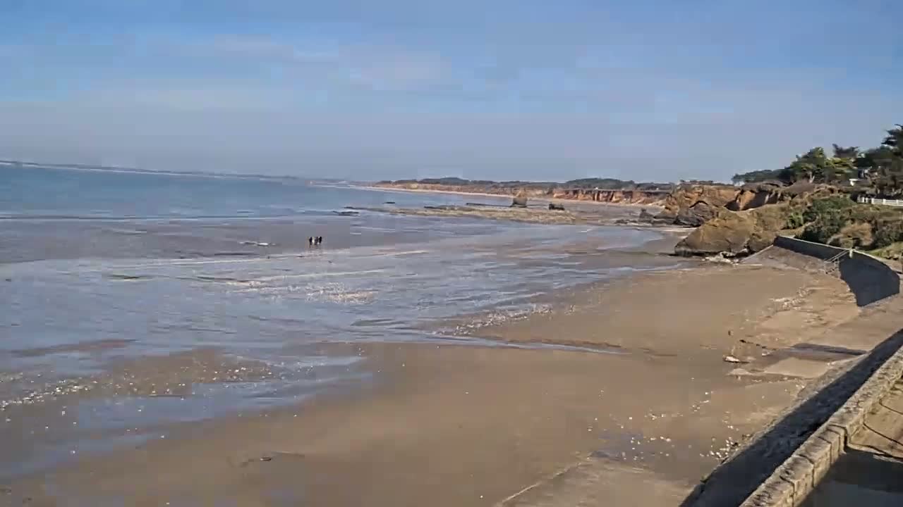 A sandy beach stretches out to a calm ocean under a clear, sunny sky, with a few people walking near the water's edge and rocky cliffs in the background.