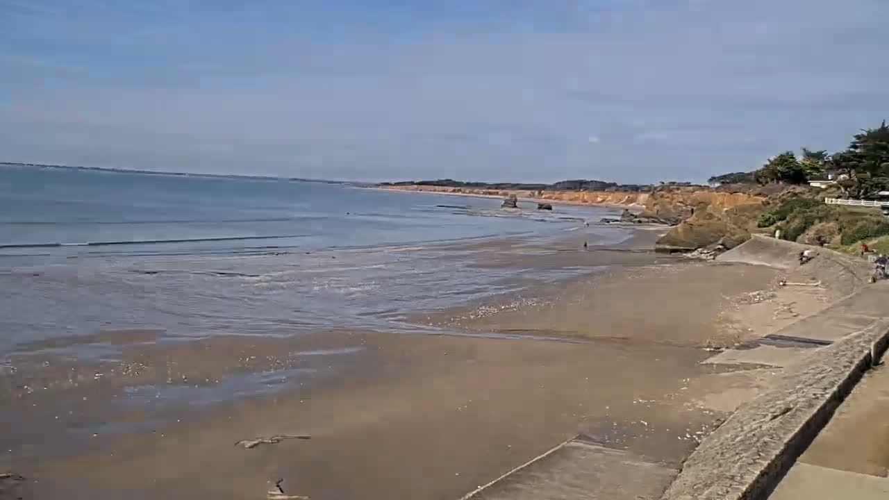 A sandy beach with calm waves stretches out to a calm ocean under a clear blue sky, bordered by a cliff with vegetation and a stone wall.