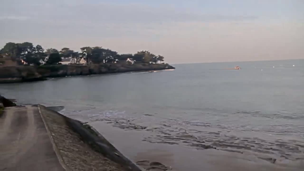 A calm, overcast day at a coastal area shows a small boat on the water and a paved road leading to a rocky shoreline with trees.