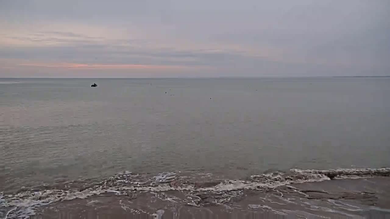 A calm sea with small waves lapping a shoreline under an overcast sky shows a distant small boat.