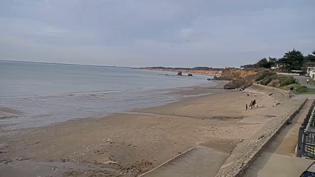 A wide sandy beach stretches along a calm, grey sea under an overcast sky, with a few people and a horse near the shore and red cliffs in the distance.