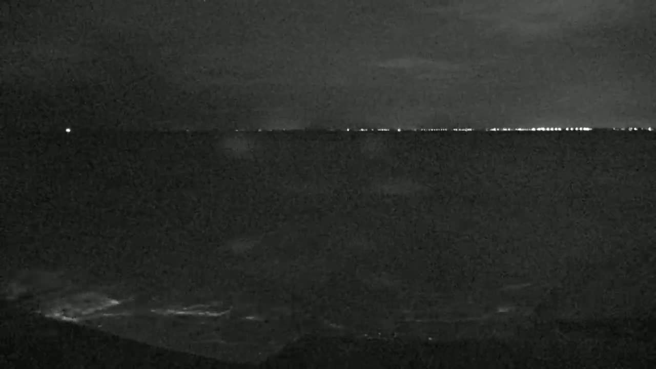 A dark, nighttime scene shows a distant line of lights across a body of water with a slightly wavy surface.