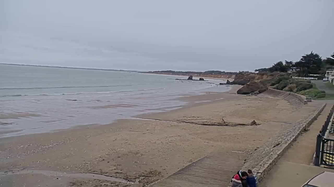 A sandy beach stretches to a calm, gray sea under an overcast sky, with a stone wall and a few people near the shore.