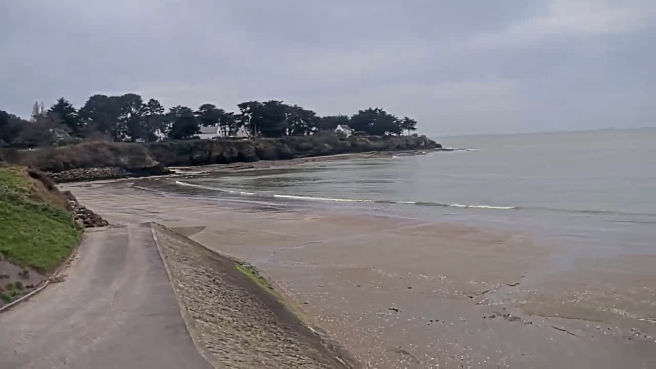 A sandy beach and calm, gray water are visible under an overcast sky, with a paved road leading down to the water's edge, bordered by green vegetation and trees on a cliff in the background.
