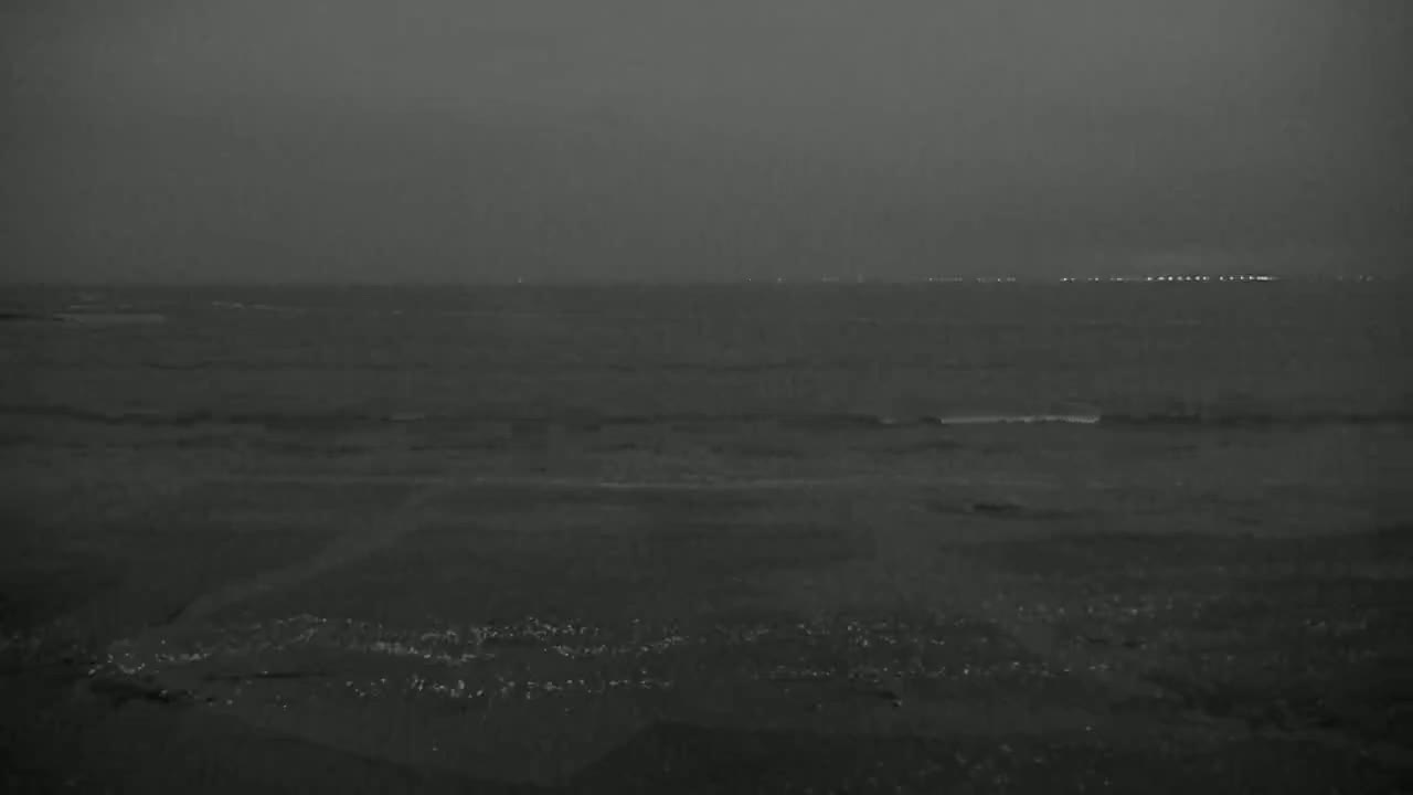 A dark, overcast sky looms over a calm, dark ocean with a distant shoreline faintly illuminated by lights.