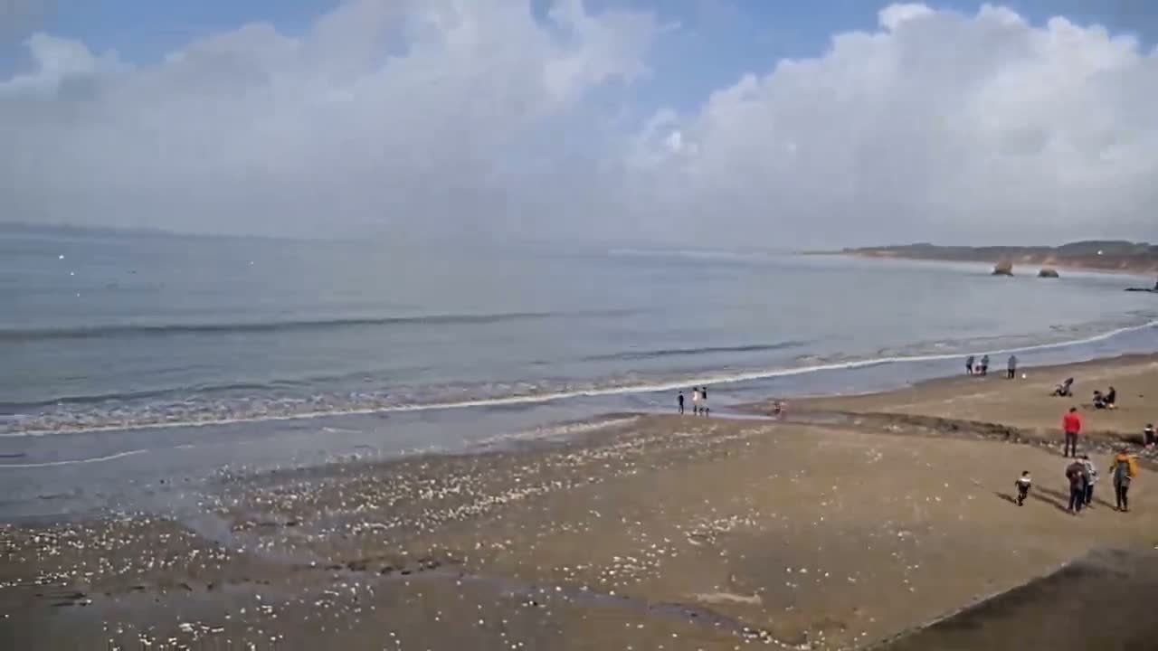 A sandy beach with a few people scattered across it and gentle waves lapping the shore under a partly cloudy sky.