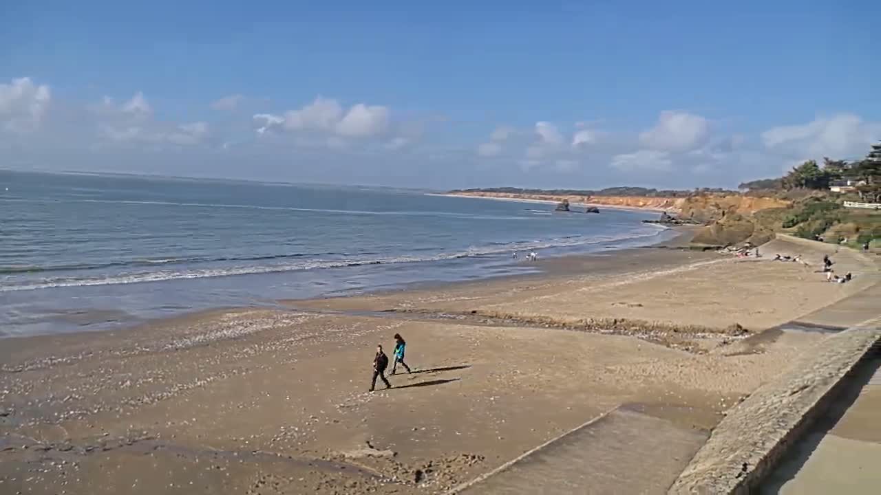 A sandy beach stretches along a calm ocean under a mostly sunny sky, with a few people walking along the shore.