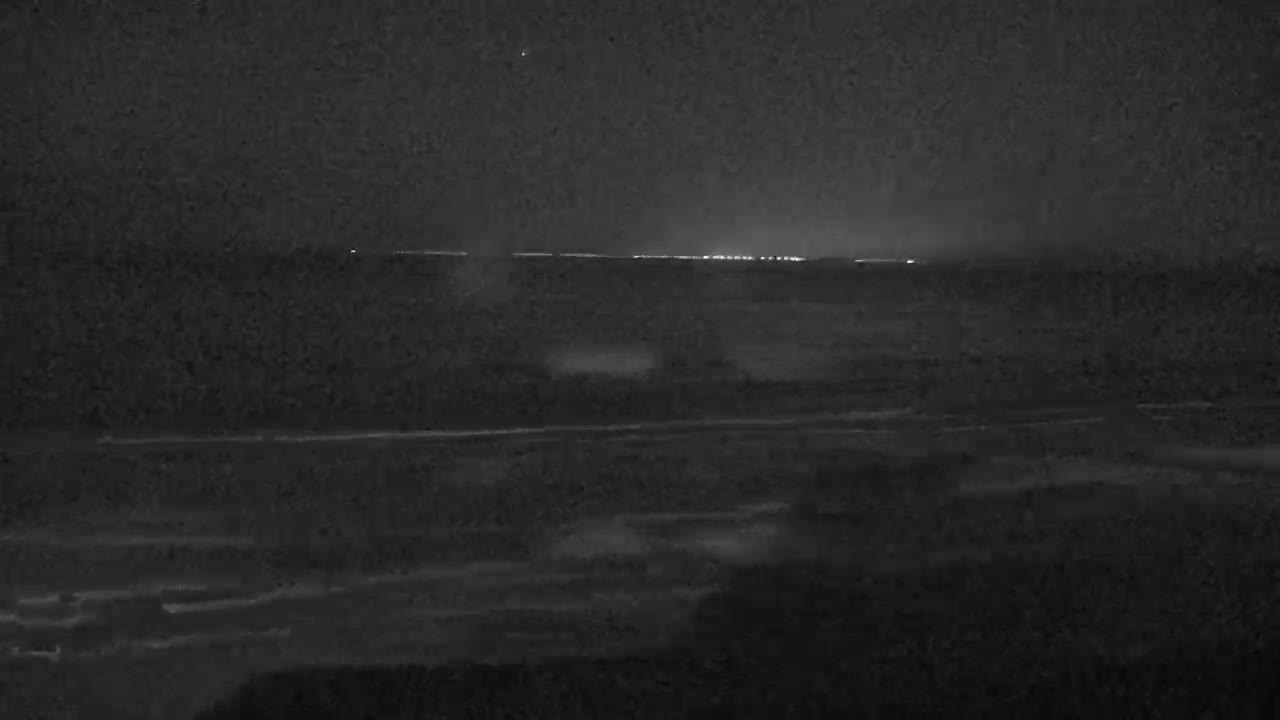 A dark, nighttime scene shows a calm ocean with a distant, faintly lit shoreline under a clear sky.