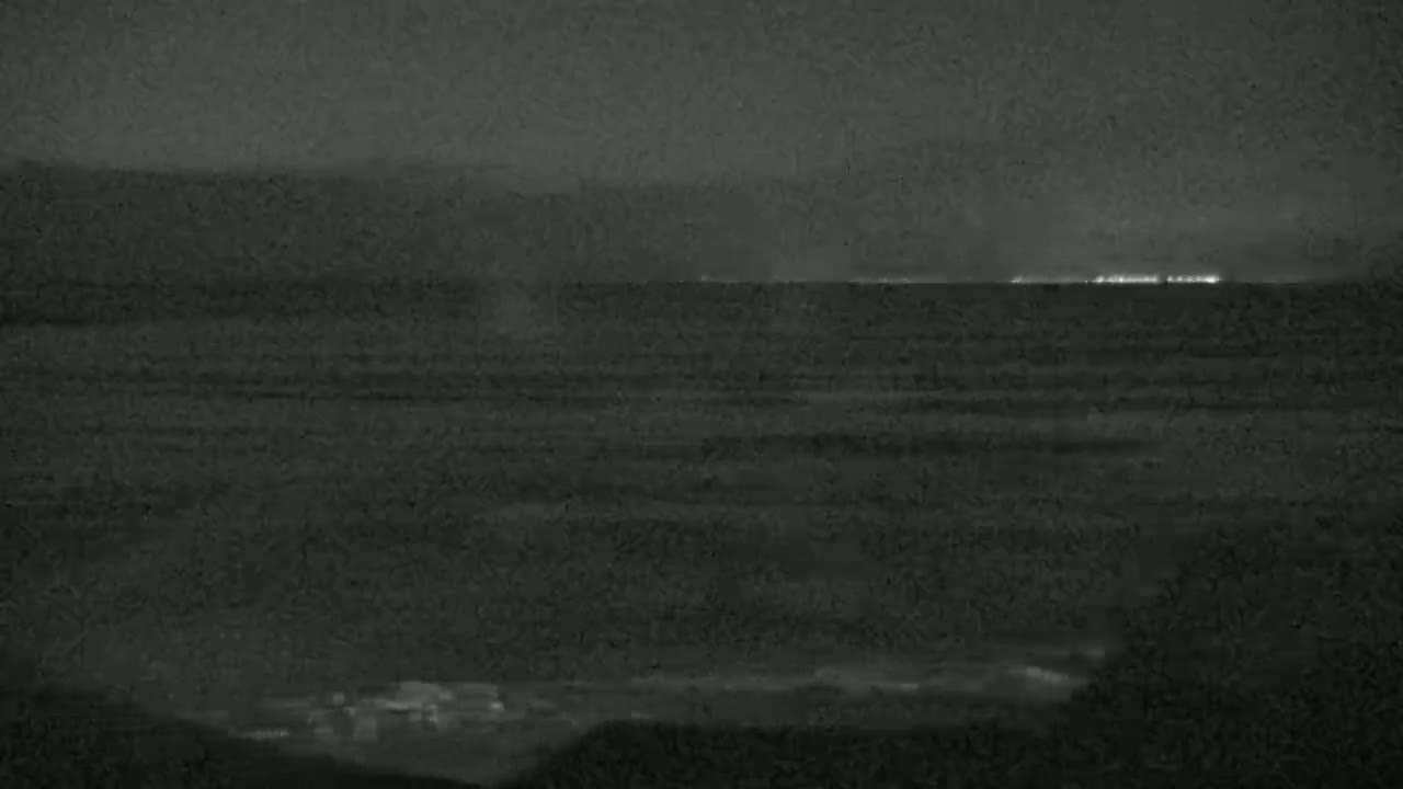 A dark, nighttime scene shows a body of water with a distant, faintly lit shoreline under an overcast sky.