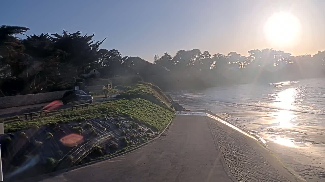 A paved path leads down to a beach where gentle waves lap the shore under a bright, sunny sky.