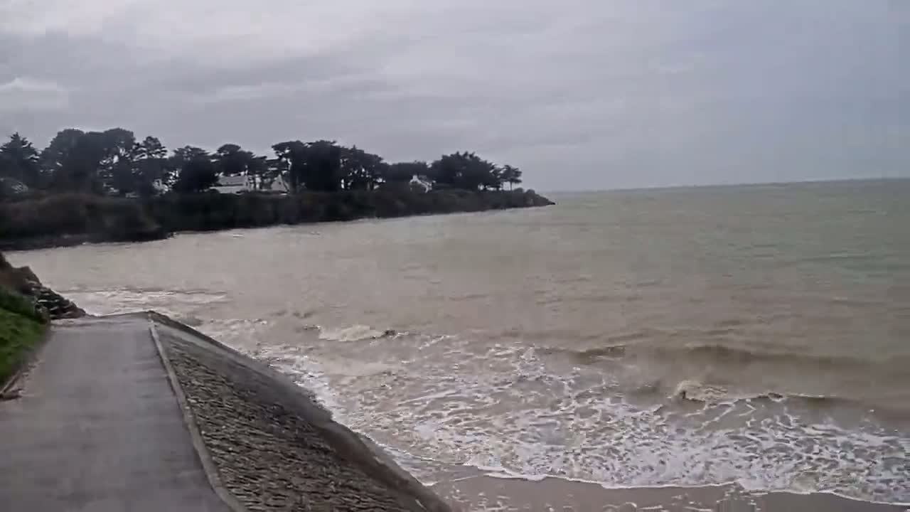 A paved path leads to a muddy, wavy ocean under an overcast sky, with trees and houses visible on a distant headland.