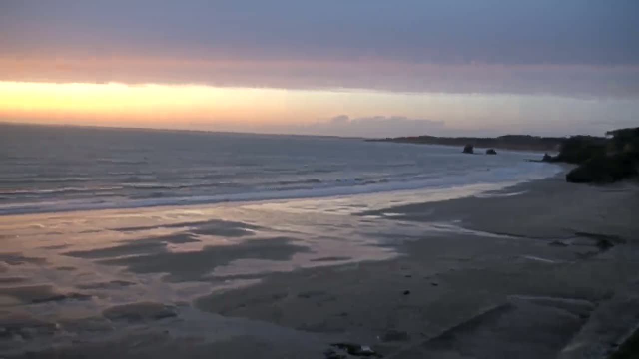 A sandy beach with small waves rolls onto the shore under a partly cloudy sunset sky.