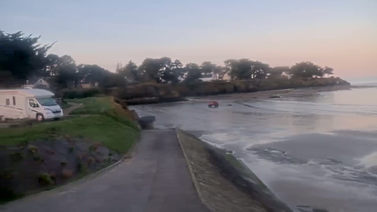 A camper van is parked near a grassy area overlooking a calm, light-brown body of water at dawn, with a small vehicle on the water's edge.