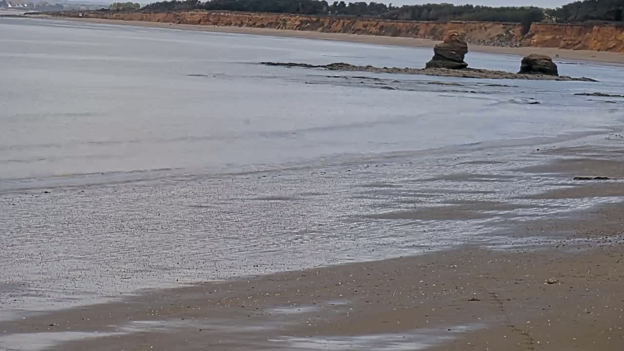 A sandy beach with gentle waves laps against the shore, featuring two rock formations jutting out from the water near a distant cliff.