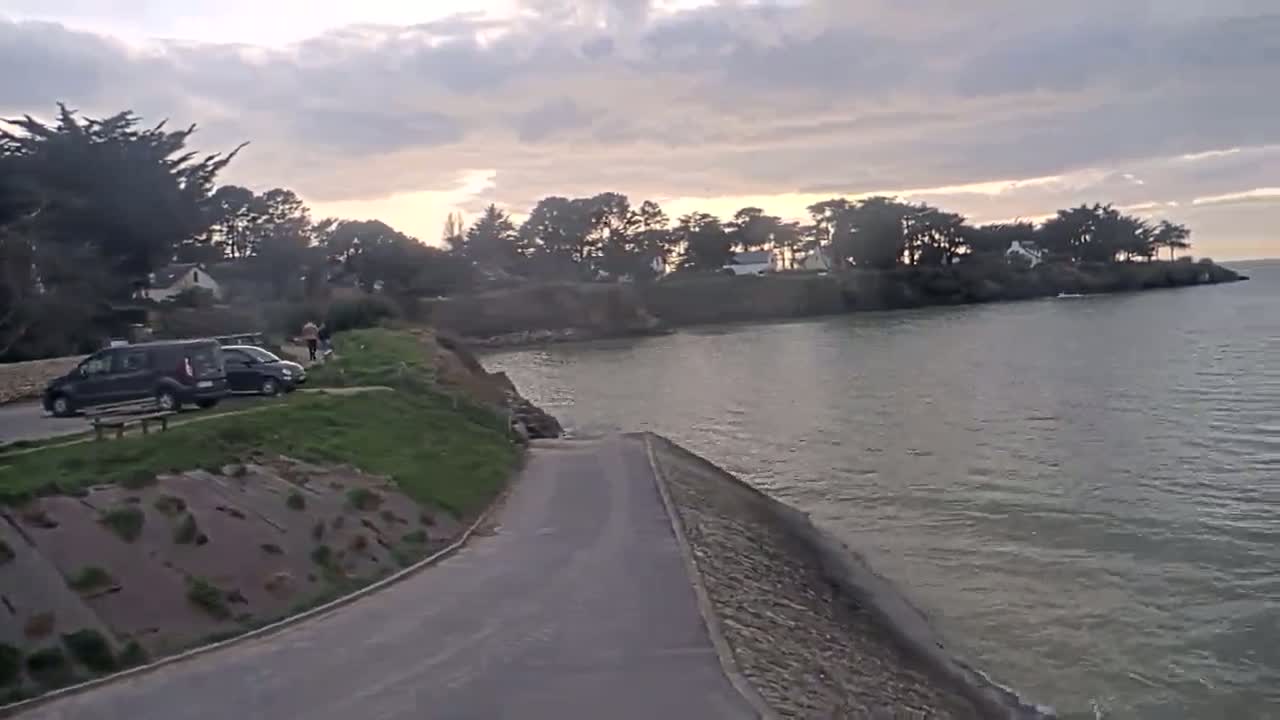 A paved road leads down to a calm body of water under a cloudy sunset sky, with parked cars and a few people visible near a shoreline with trees and houses.