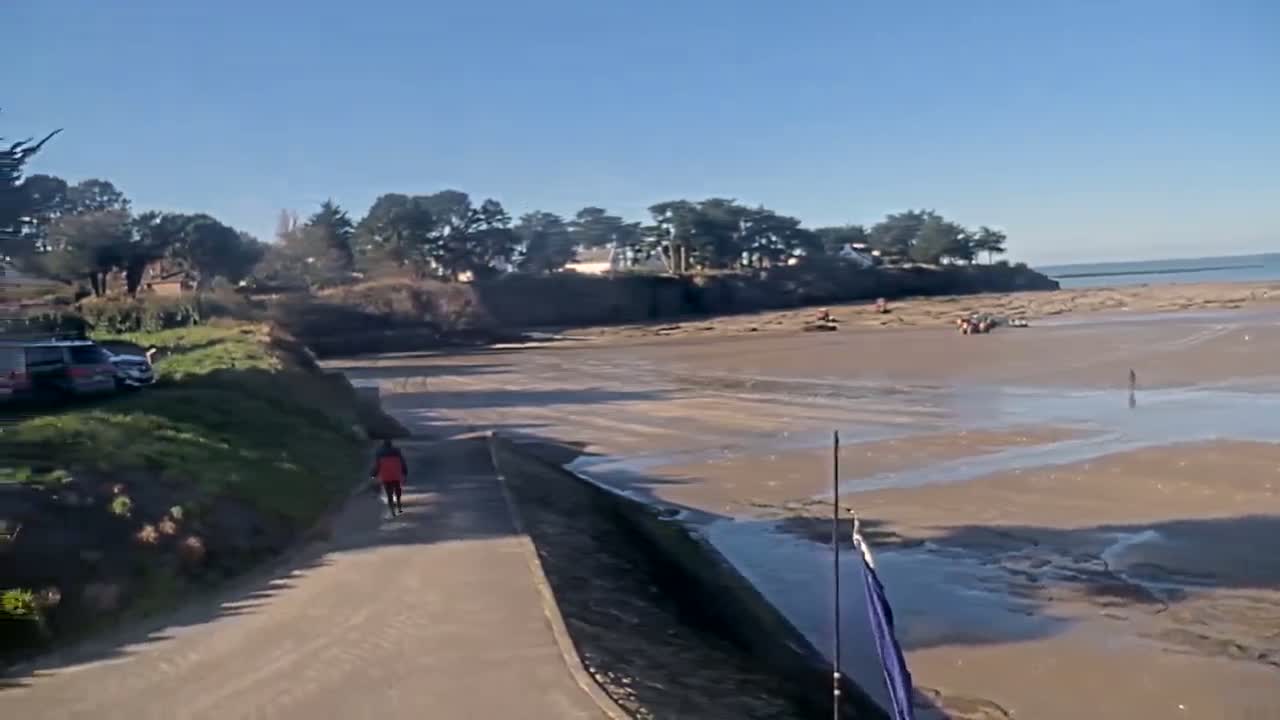 A person walks along a paved path overlooking a wide, mostly flat, muddy beach under a clear, sunny sky.