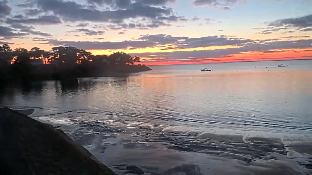 A tranquil sunrise paints the sky with vibrant orange and pink hues over a calm bay, where a few small boats are visible near a silhouetted shoreline with trees.
