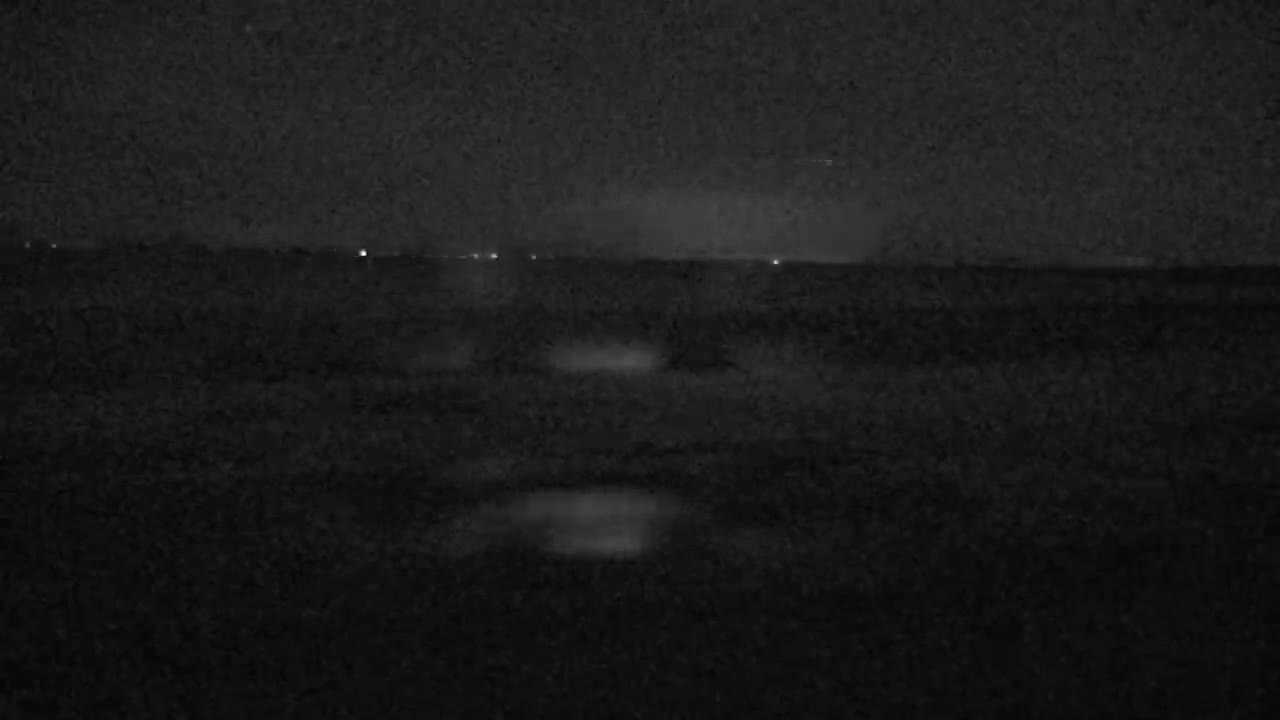 A dark, nighttime outdoor scene shows a flat, seemingly empty landscape with a few distant lights and faint reflections on a wet surface.