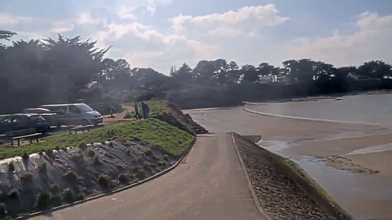 A paved path leads down to a sandy beach with calm water under a partly cloudy sky, where several people stand near parked cars and a van.
