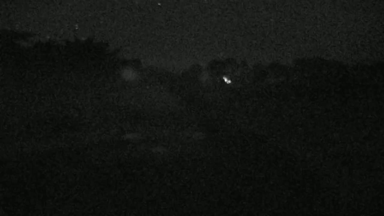 A low-light, nighttime image shows a dark, possibly wooded area with a few faint lights visible in the distance.