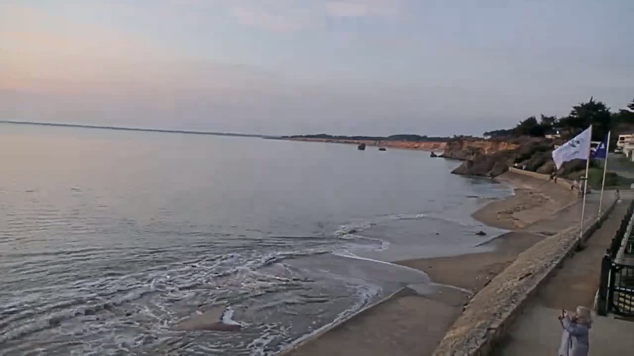 A calm, pale-grey ocean laps a sandy beach next to a low, rocky cliff under a dusky sky.