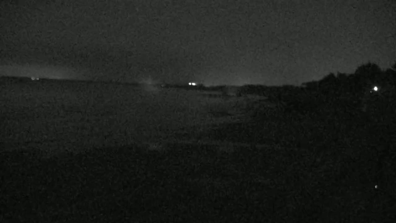 A dark, nighttime scene shows a flat expanse of land with a few distant lights under a cloudy sky.