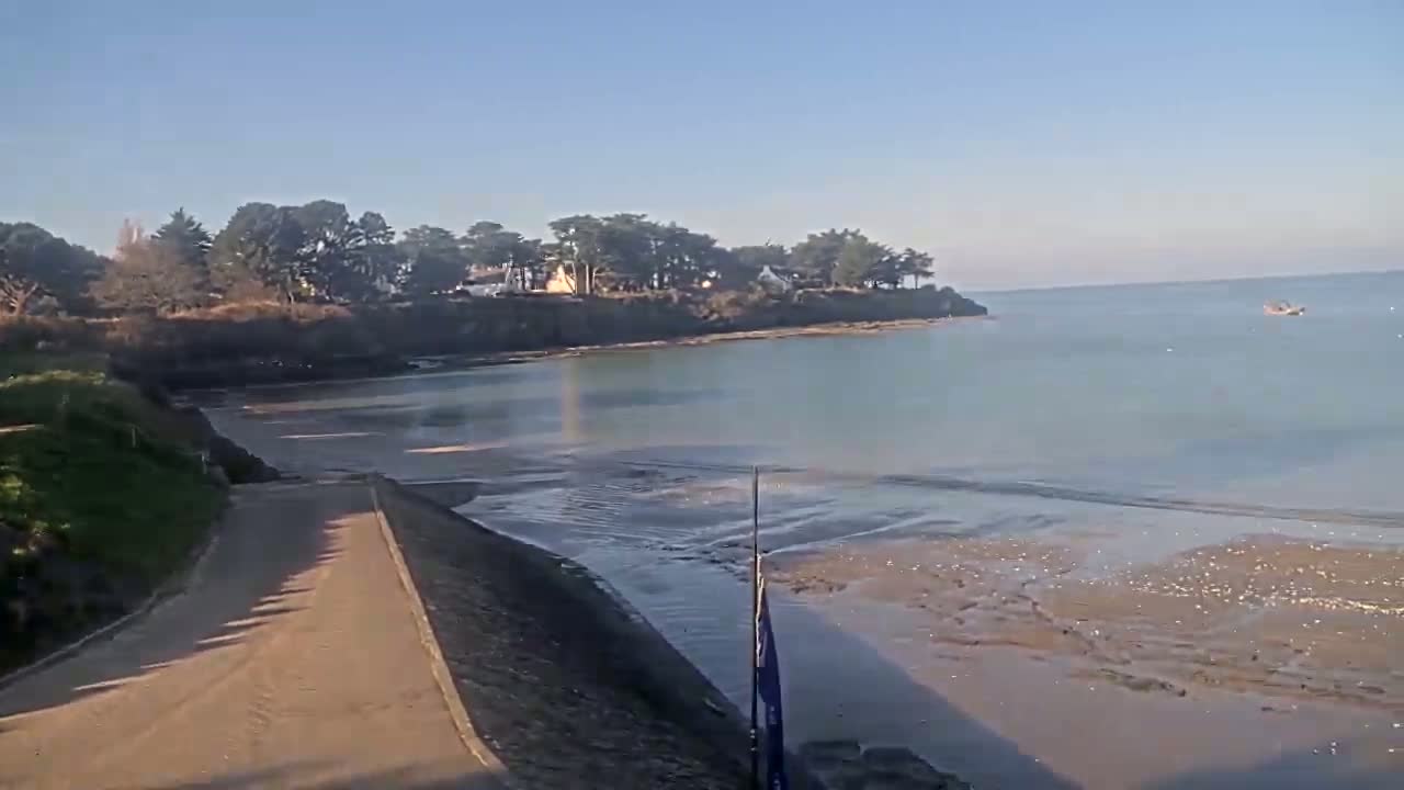 A sandy path leads to a calm bay under a clear blue sky, with a small boat visible in the distance and a line of trees on a small cliff in the background.