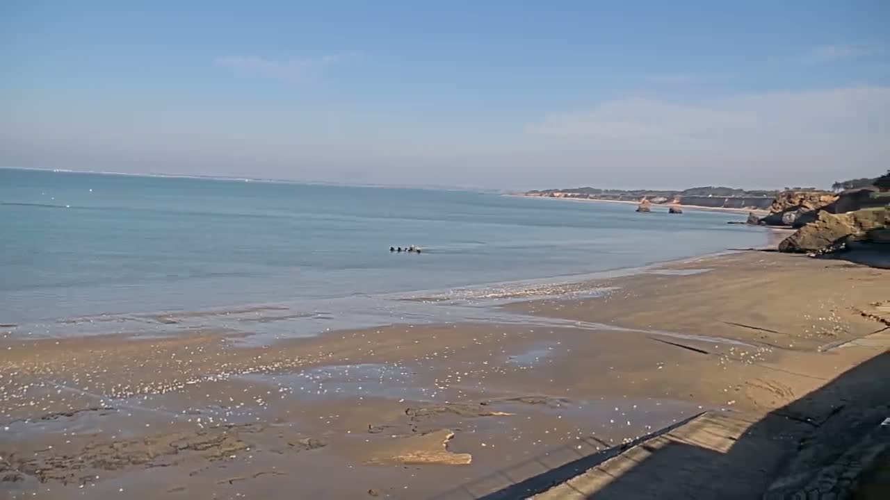 A sandy beach stretches along a calm, blue ocean under a clear sky, with small rock formations and cliffs visible in the distance.