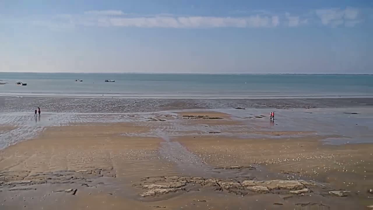 A wide sandy beach, mostly exposed at low tide, stretches to a calm, light-blue sea under a mostly clear sky with a few wispy clouds, and a few small figures are walking on the wet sand.