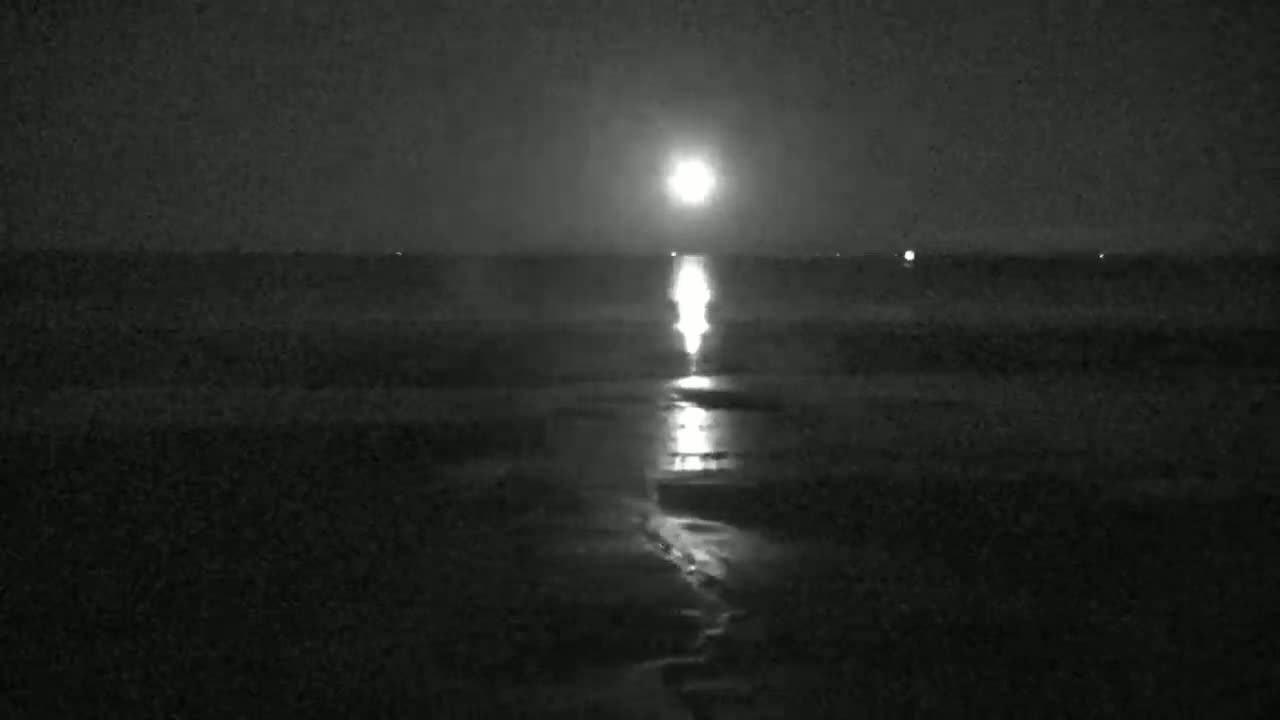 A bright moon shines over a dark body of water at night, its reflection creating a luminous path across the surface.