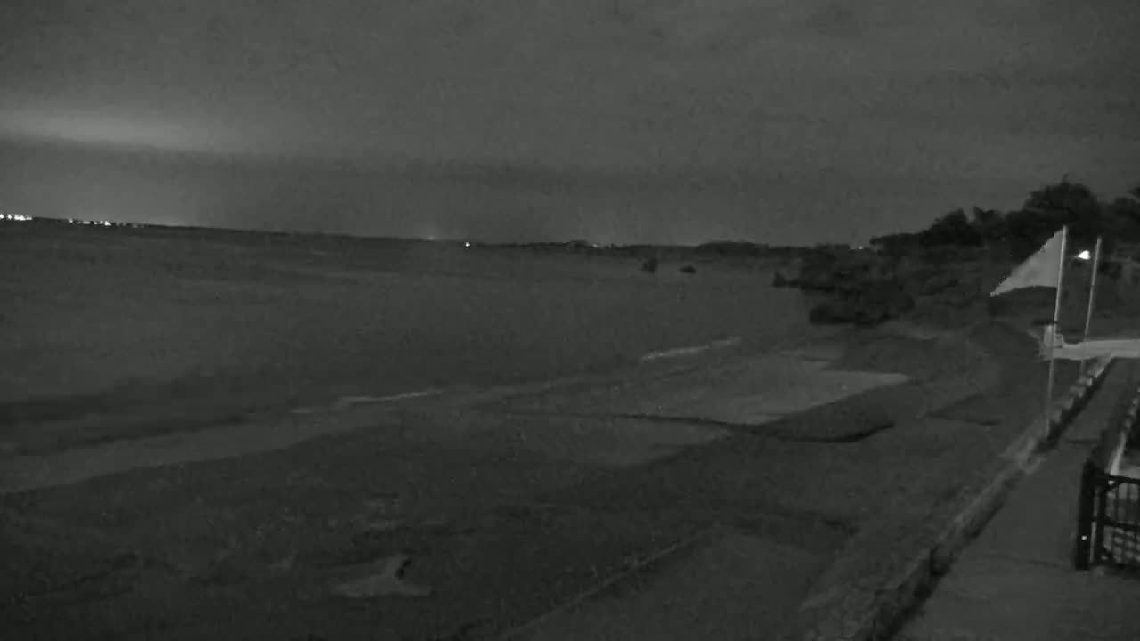 A dark, nighttime beach scene shows a mostly flat, sandy shore under a cloudy sky, with a few lights visible in the distance.