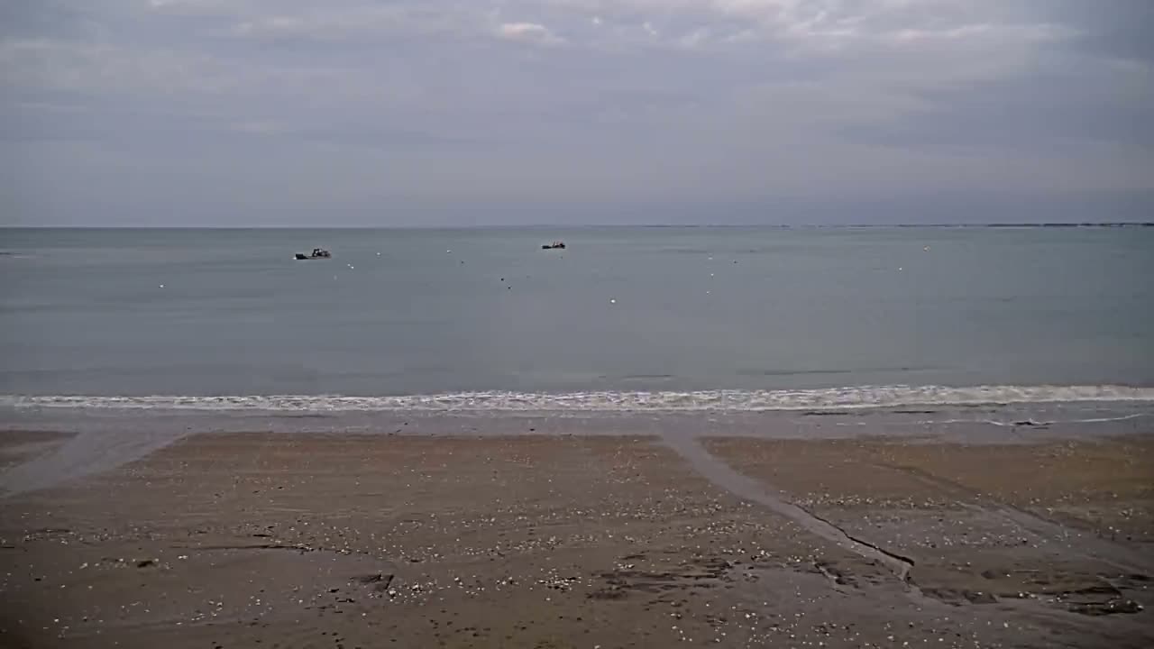 A sandy beach with small waves gently lapping the shore under a cloudy sky, with a few small boats visible in the distance on calm water.