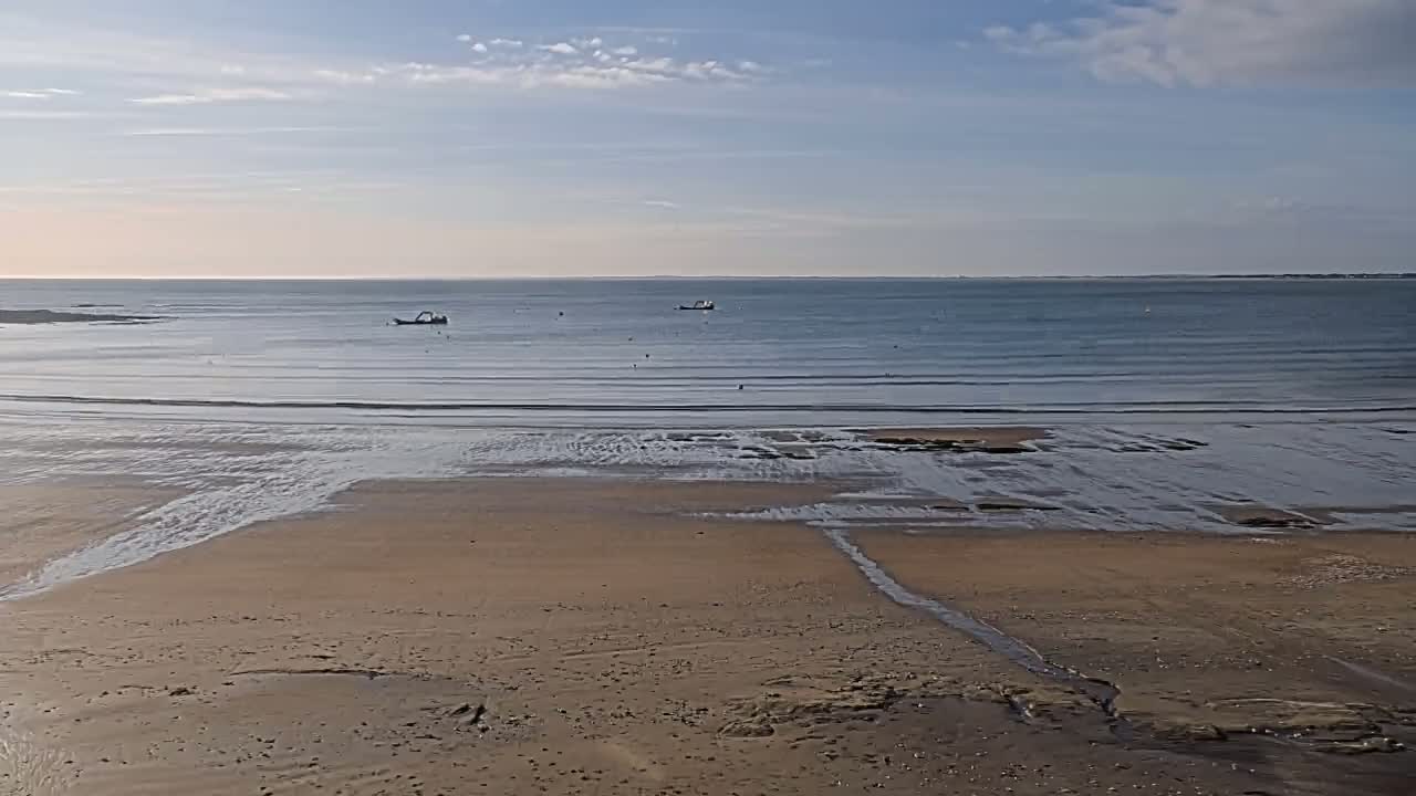 A sandy beach stretches to a calm sea under a mostly clear sky, with a few small boats visible on the water.