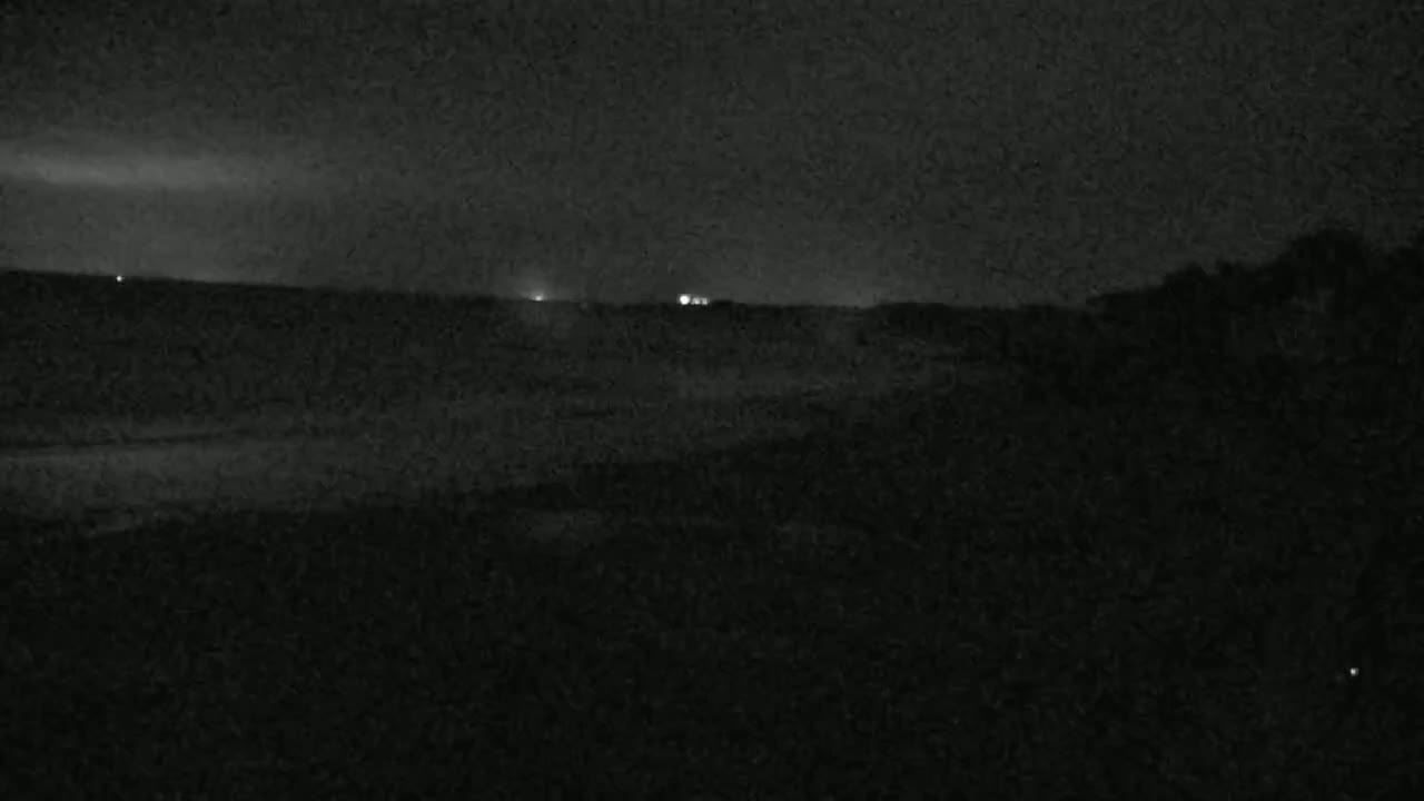 A dark, nighttime landscape shows a flat horizon with a few distant lights under a cloudy sky.