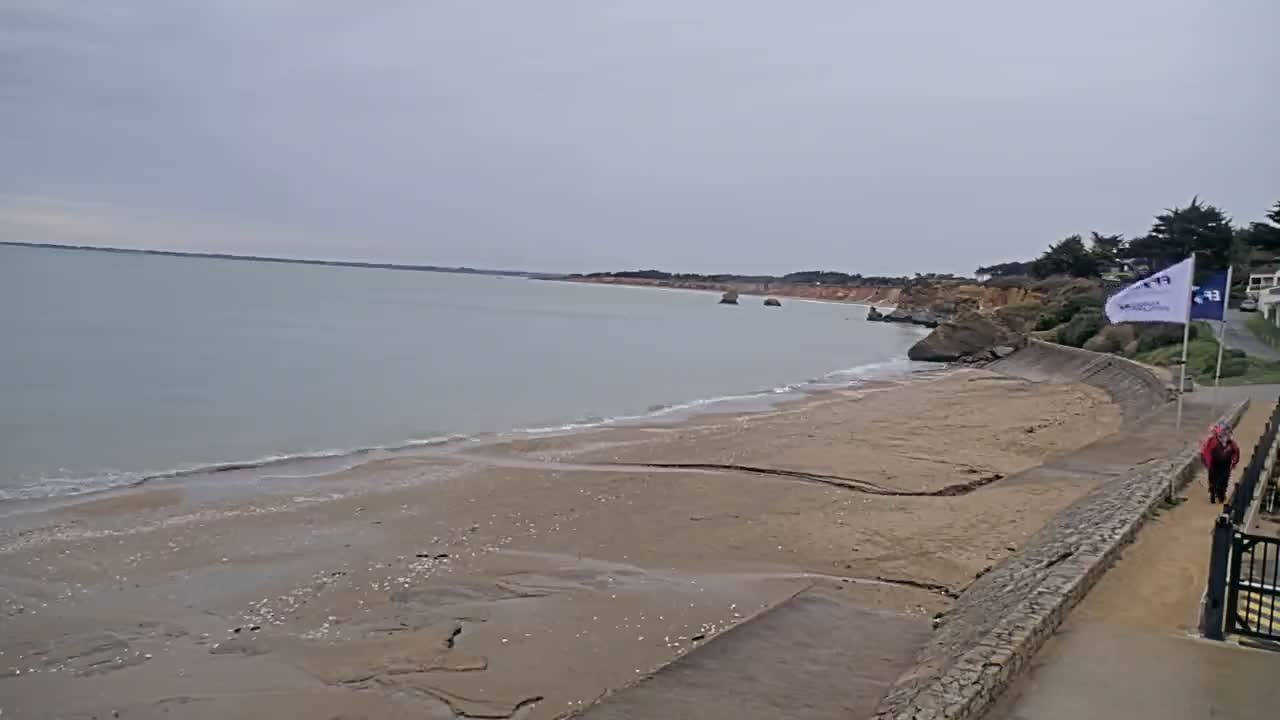 A sandy beach stretches along a calm, gray sea under an overcast sky, with a seawall and walkway bordering the beach.