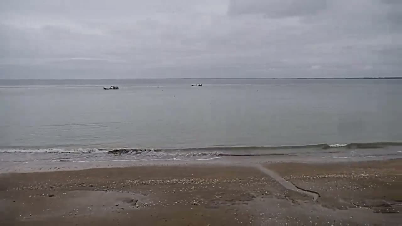 A sandy beach meets a calm, gray sea under an overcast sky, with two small boats visible in the distance.