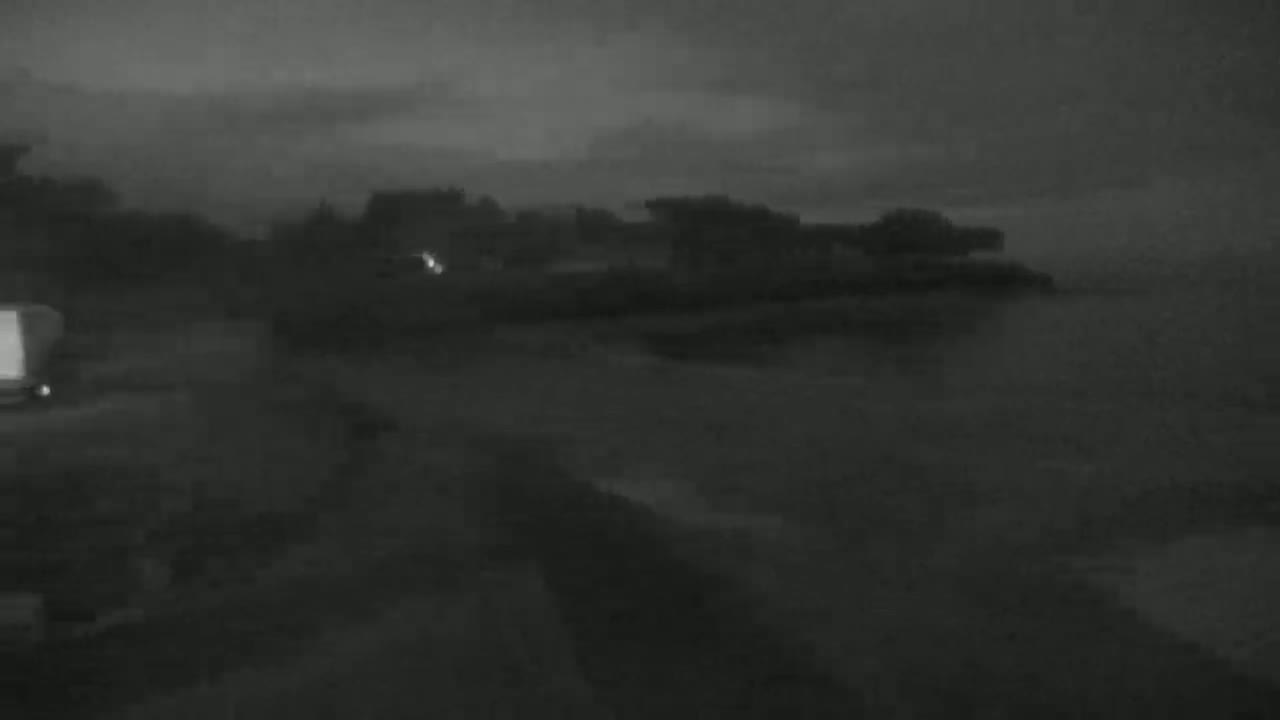 A dark, overcast night scene shows a body of water next to a shoreline with some vegetation, and a vehicle is visible on the left.