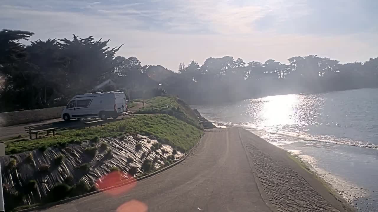 A camper van is parked near a paved road leading to a calm body of water under a partly sunny sky.