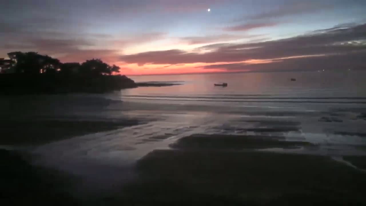 A pink and orange sunset over a calm ocean with a few small boats and a dark shoreline with trees.