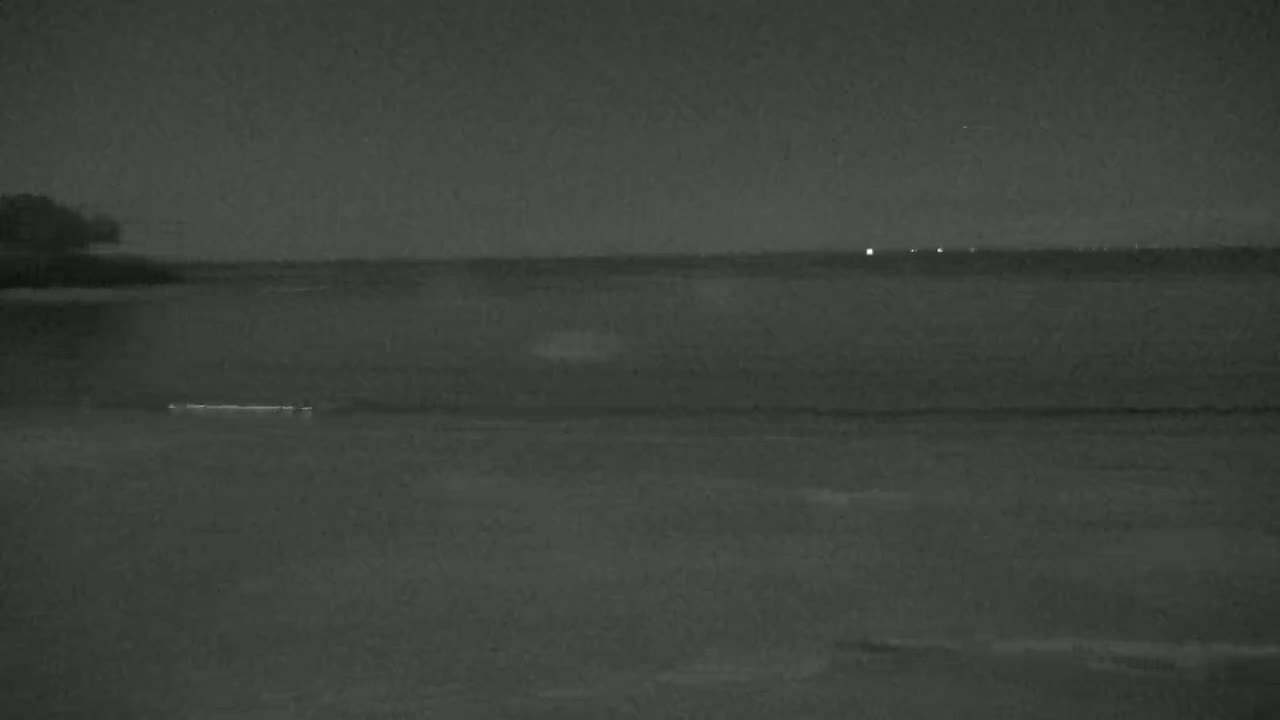 A dark, nighttime view of a calm body of water under a cloudy sky, with faint lights visible on the horizon.