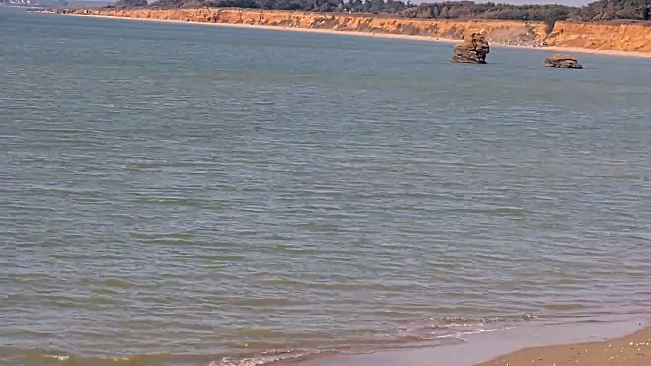 Calm, light-grey water laps a sandy shore near a distant, sandy cliff line with two small rock formations in the water.