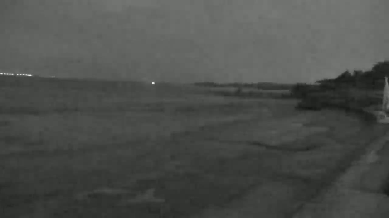 A dark, overcast night scene shows a flat expanse of land, possibly a field or beach, with some indistinct vegetation or structures in the background.