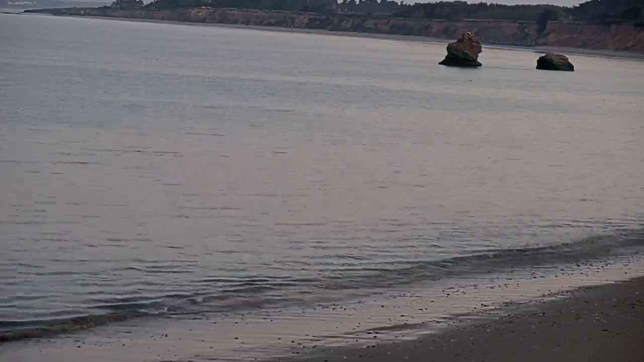 A calm, gray ocean with two small rock formations offshore and a sandy beach in the foreground.