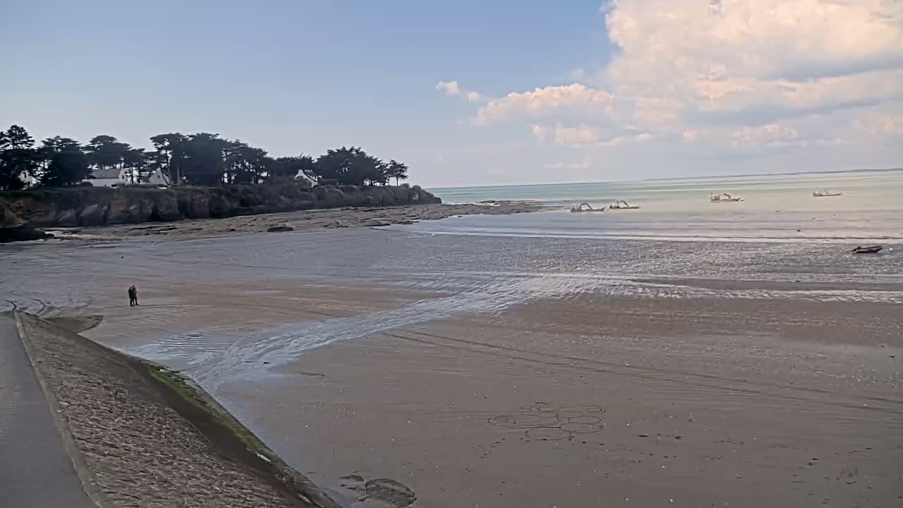 A mostly sunny day shows a sandy beach with several boats in the calm water, a few people walking on the shore, and a line of trees and houses on a cliff in the background.