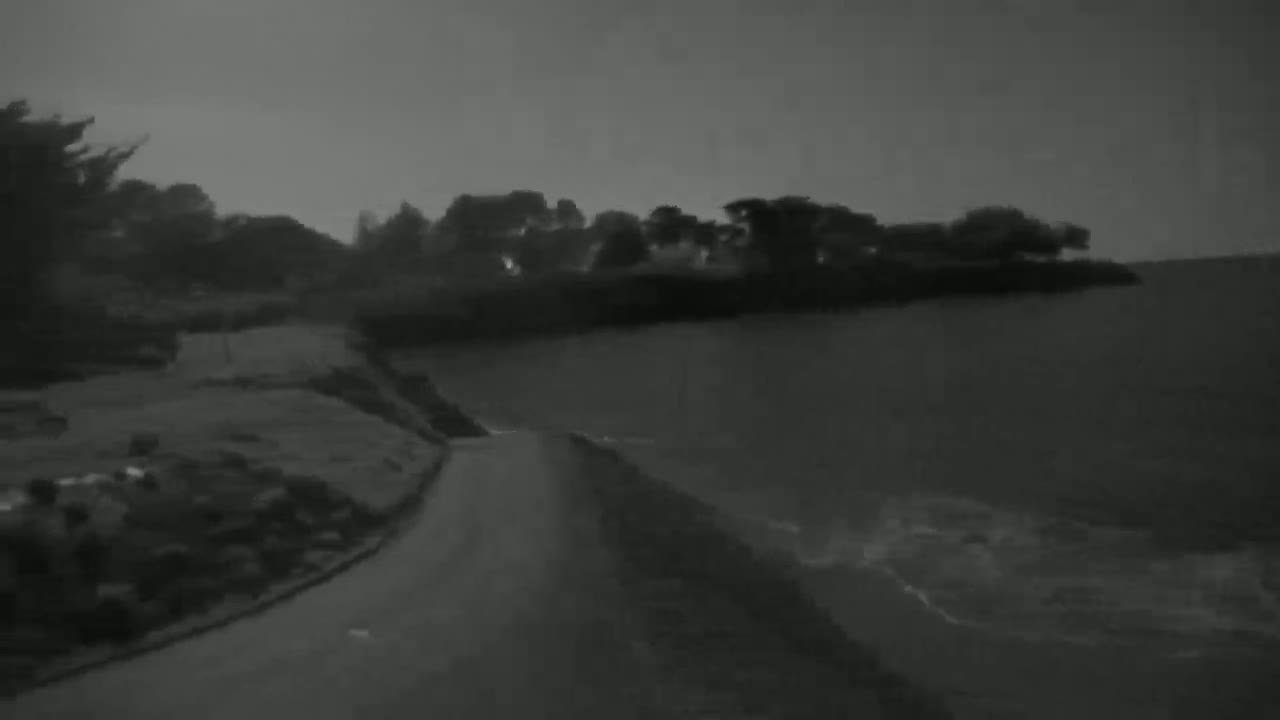 A dark gray road curves alongside a body of water under a dim, overcast sky, leading toward a shoreline lined with trees.
