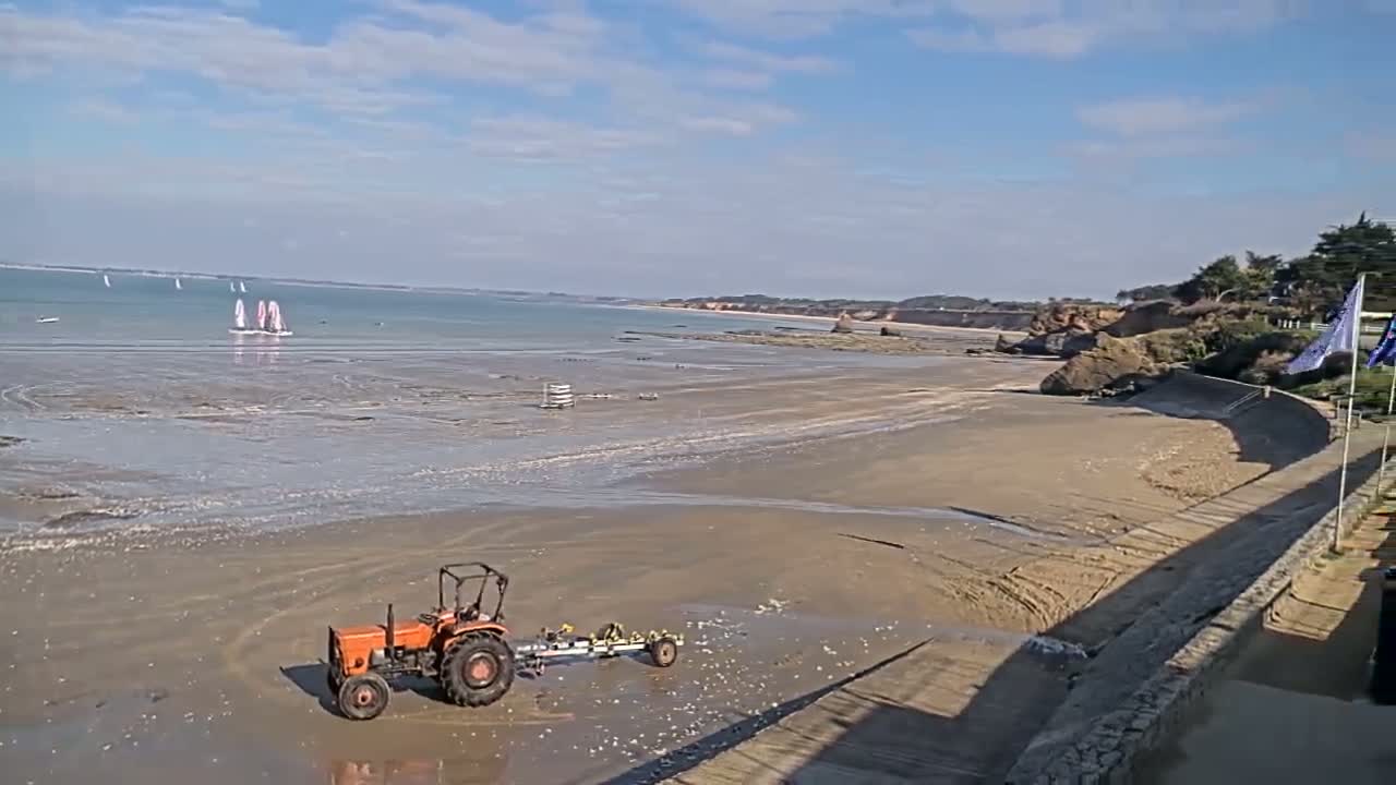 An orange tractor pulling a small trailer sits on a sandy beach near calm ocean water under a mostly sunny sky, with sailboats visible in the distance.