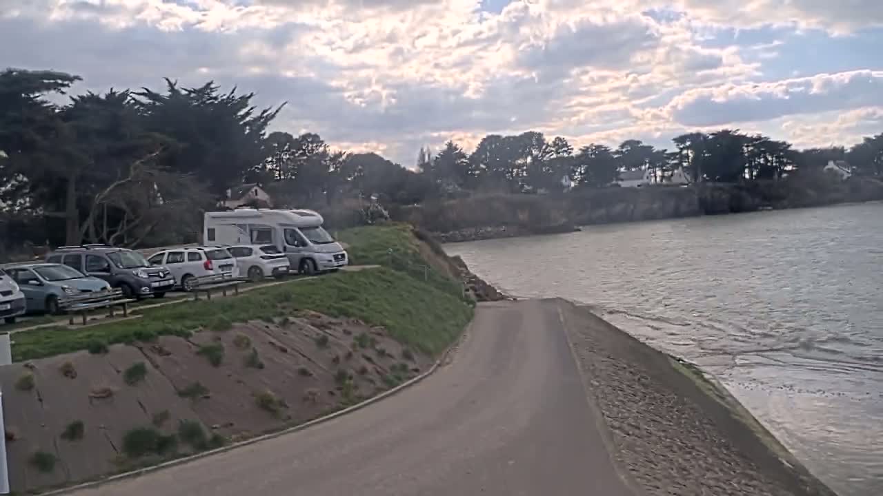 A paved road curves down to a calm body of water, next to a grassy area where several cars and a campervan are parked under a partly cloudy sky.