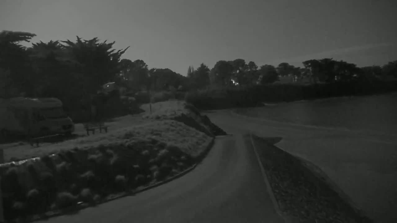 A dark, nighttime scene shows a paved road curving past a grassy area with shrubs, a campervan parked nearby, and trees in the background under a mostly clear sky.